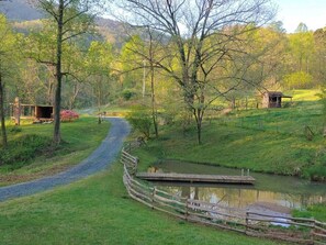 Un paisaje rural con un camino de grava, valla de madera y una pequeña construcción de madera cerca de un estanque.