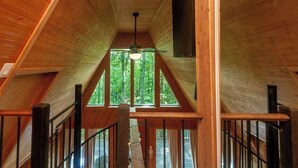 A wooden interior with a sloped ceiling, a ceiling fan, and a view of a forest through large windows. - Firepit + Mountain Views | A-Frame in Fairview | Honeycomb Hideaway by AvantStay (Fairview)