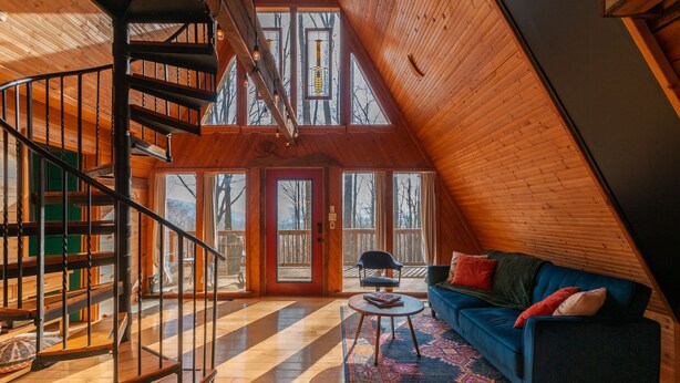A spacious loft with a wooden interior, a spiral staircase, and large windows offering views of the outdoors. - Firepit + Mountain Views | A-Frame in Fairview | Honeycomb Hideaway by AvantStay (Fairview)