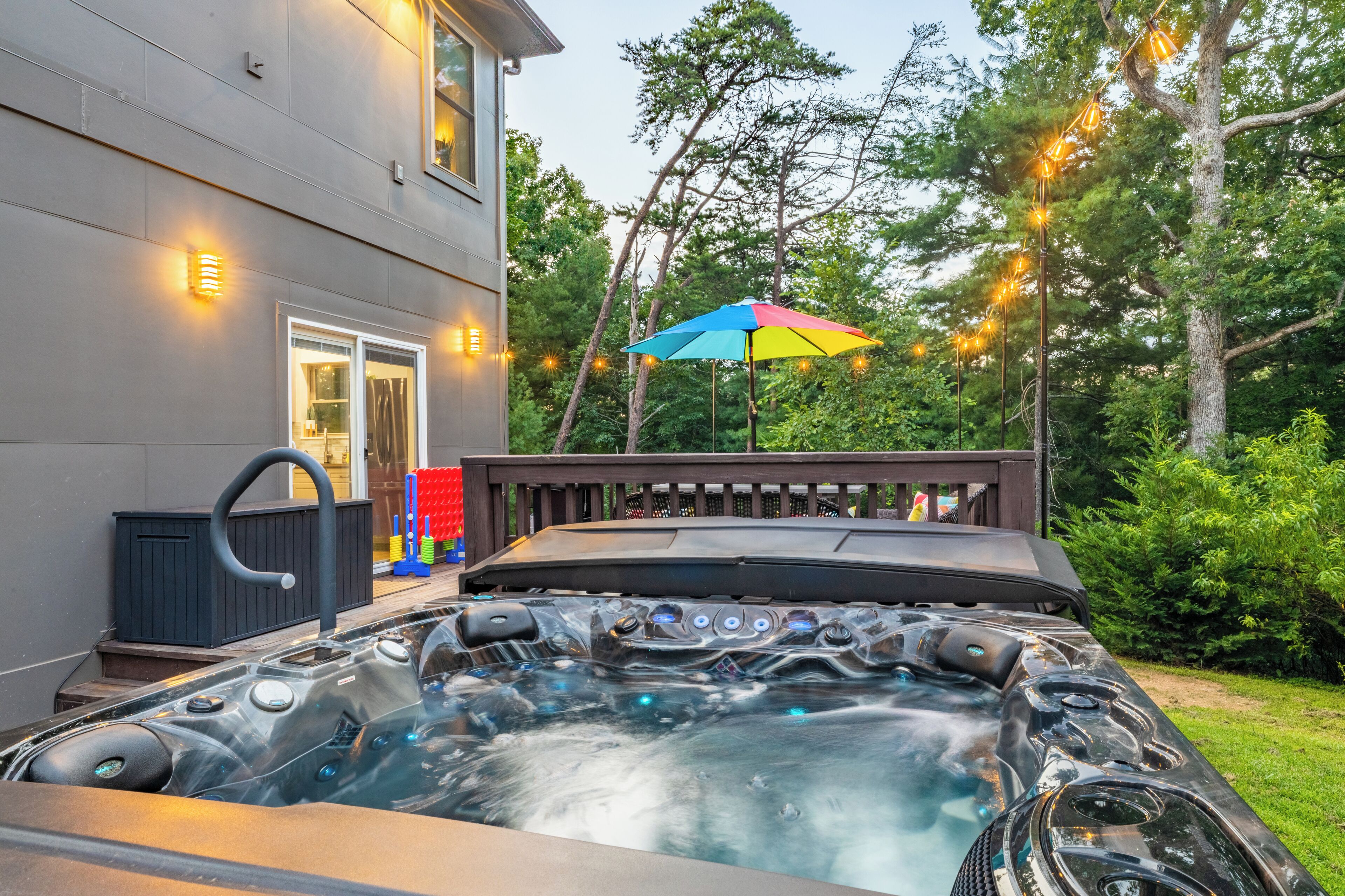 A hot tub with a wooden deck and a colorful umbrella, surrounded by trees and string lights.