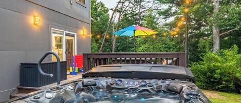 A hot tub with a wooden deck and a colorful umbrella, surrounded by trees and string lights.
