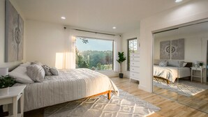 A modern bedroom with a large bed, a dresser, a mirror, and a view of a forest through a large window. - Hot Tub, Fire Pit, Mountain Views, 10min to AVL |  Modern Escape by Avanstay (Asheville)