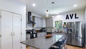 A modern kitchen with white cabinets, a central island with bar stools, a stainless steel refrigerator, and a large window providing natural light. - Hot Tub, Fire Pit, Mountain Views, 10min to AVL |  Modern Escape by Avanstay (Asheville)