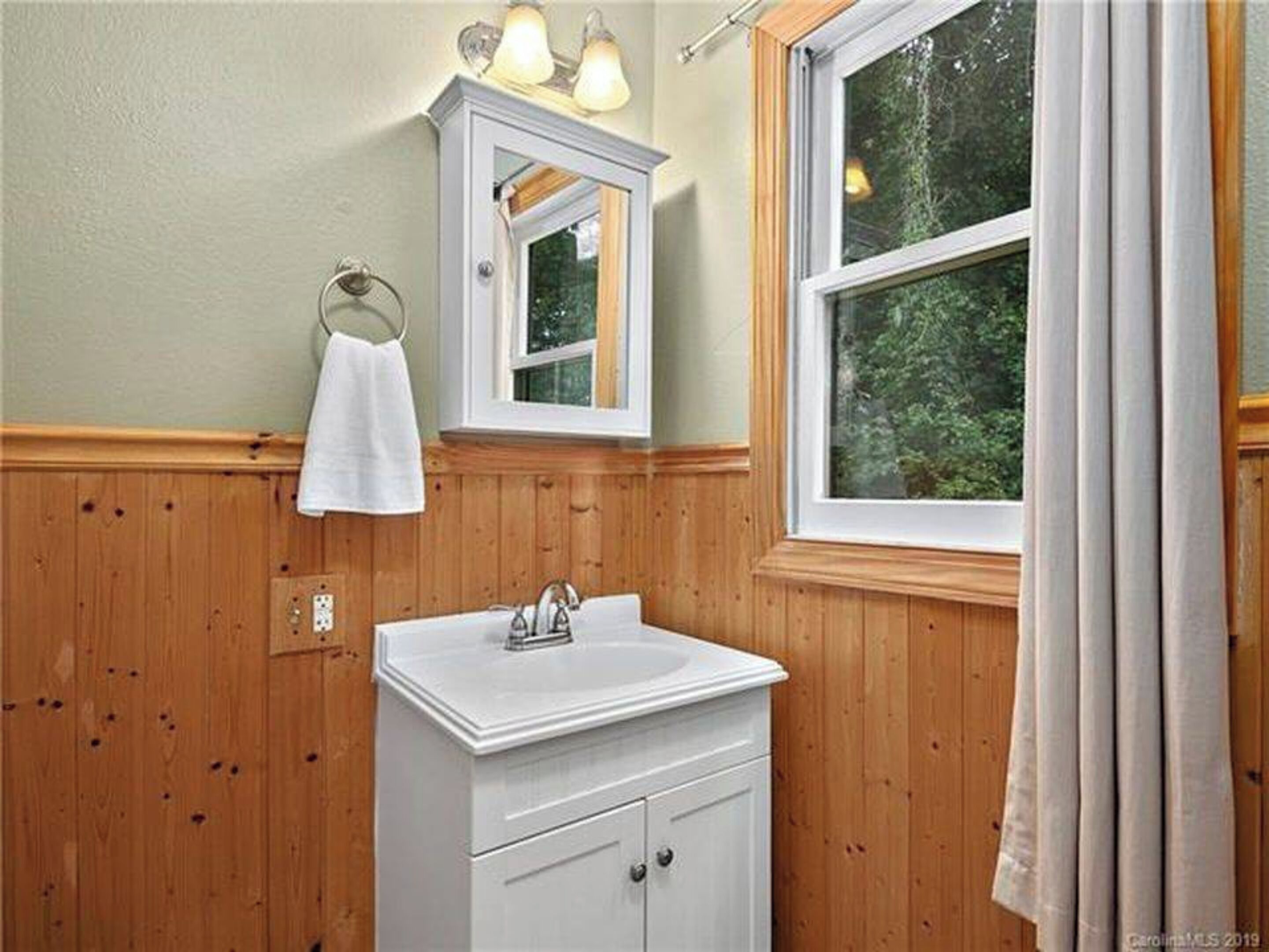 Un baño con paneles de madera, un lavabo blanco, un espejo y una ventana con cortinas.