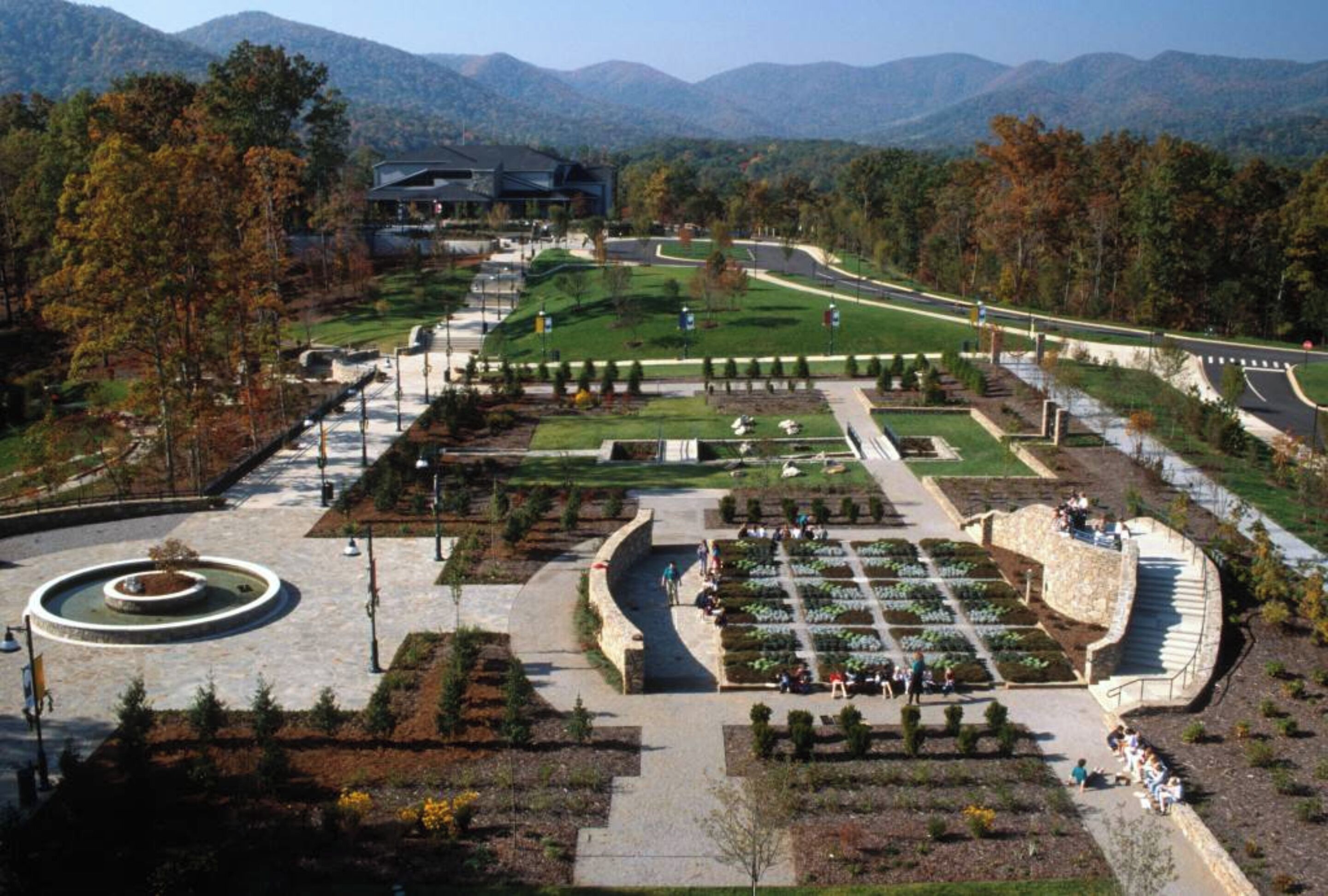 A landscaped garden with pathways, a central fountain, and a building in the background.