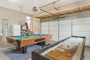 Une table de billard, avec une table de jeu de palet adjacente, un projecteur fixé au plafond et, au fond, une porte de garage.