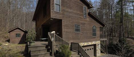 A two-story wooden cabin with a stone foundation, a front porch, and a small attached shed.