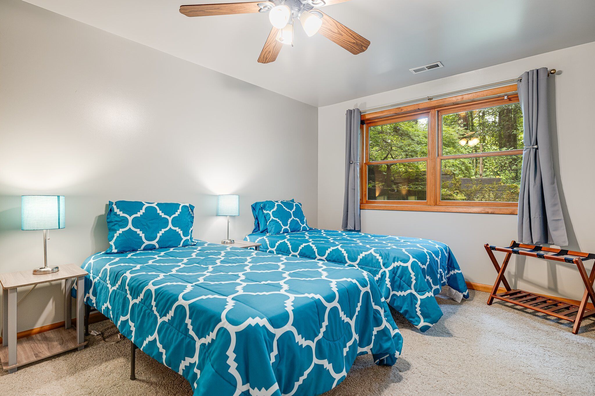 A bedroom with two beds, a ceiling fan, a window with curtains, and a small table with a lamp.