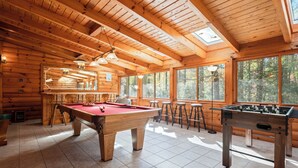 A wooden cabin interior with a pool table, foosball table, and large windows offering views of the forest.