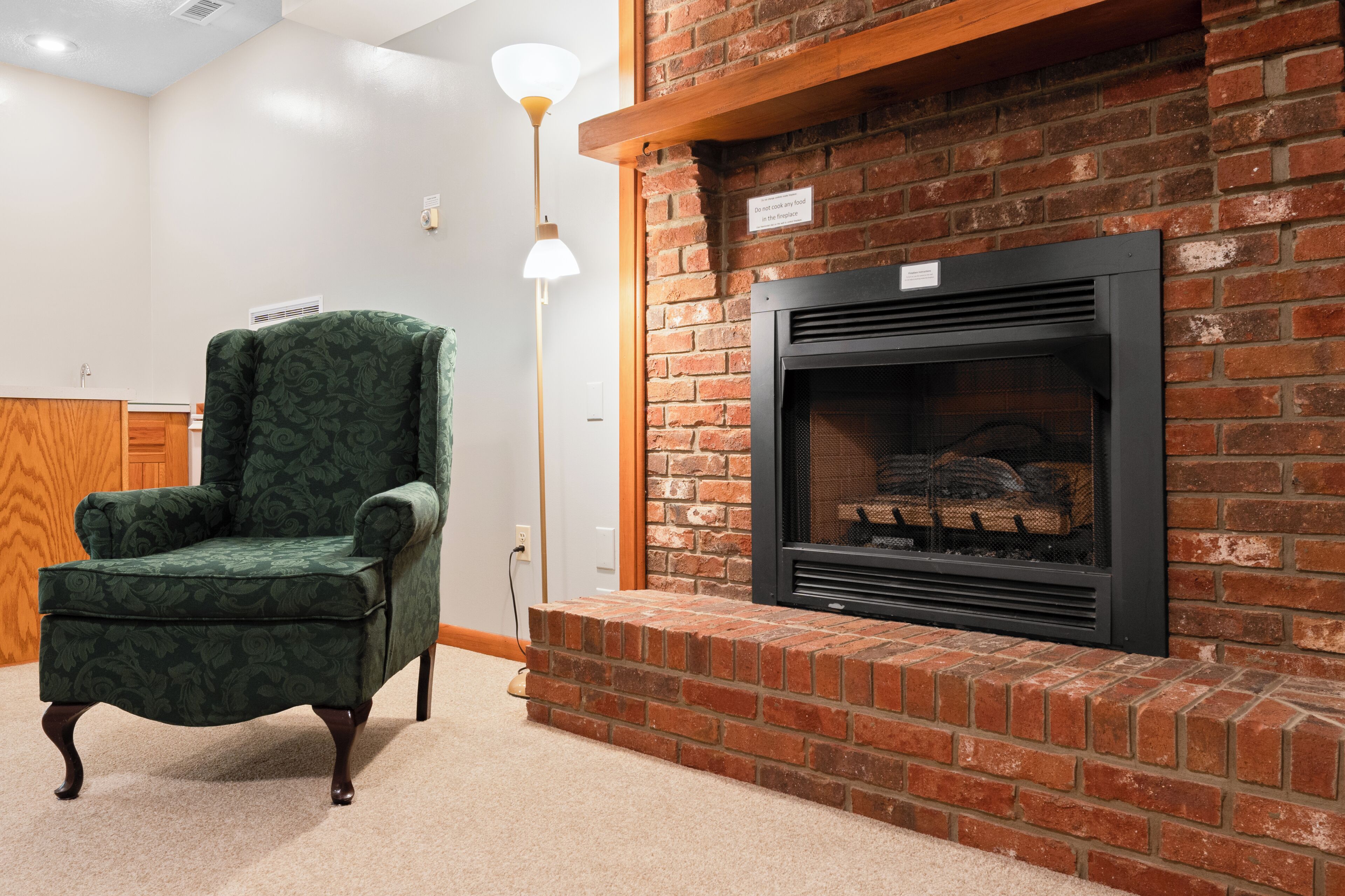Una habitación con chimenea de ladrillo, un sillón tapizado en verde y una lámpara de pie.
