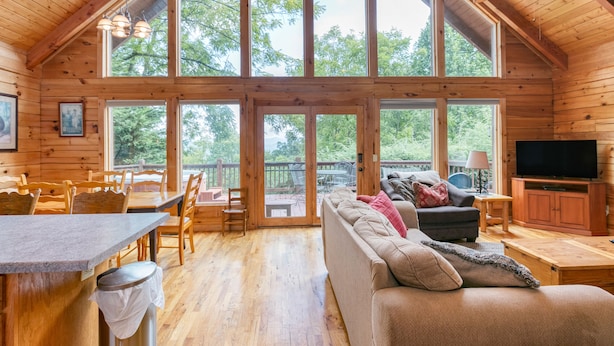 A spacious wooden cabin with a large dining area, a cozy living room with a sofa and TV, and large windows offering a view of the outdoors. - Incredible View, Hot Tub, Fire Pit, Grill & Foosball | Mountain Gem by AvantStay (Waynesville)