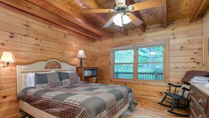 A wooden cabin bedroom with a bed, a rocking chair, a dresser, and a window with a view of trees. - Incredible View, Hot Tub, Fire Pit, Grill & Foosball | Mountain Gem by AvantStay (Waynesville)