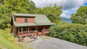 A rustic log cabin with a porch and a small deck, surrounded by lush greenery and mountains in the background. - Incredible View, Hot Tub, Fire Pit, Grill & Foosball | Mountain Gem by AvantStay (Waynesville)