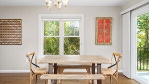 A dining area with a wooden table and chairs, a chandelier, and a large window with a view of greenery. - Little Sycamore by AvantStay | Premier Location + Patio | Sycamore Cove Gem (Asheville)