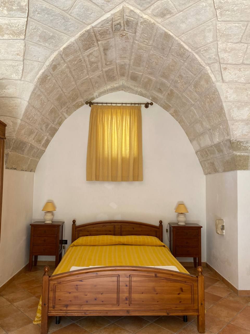 Une chambre avec un plafond en forme d’arc en pierre, un lit en bois recouvert d’une couverture jaune, deux tables de chevet avec des lampes et une fenêtre avec un rideau.