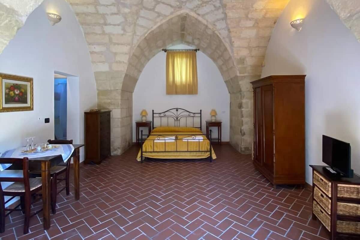 Un dormitorio con suelo de ladrillo, una cama con colcha amarilla, un armario de madera, un televisor sobre un mueble de madera y un techo con arco de piedra.