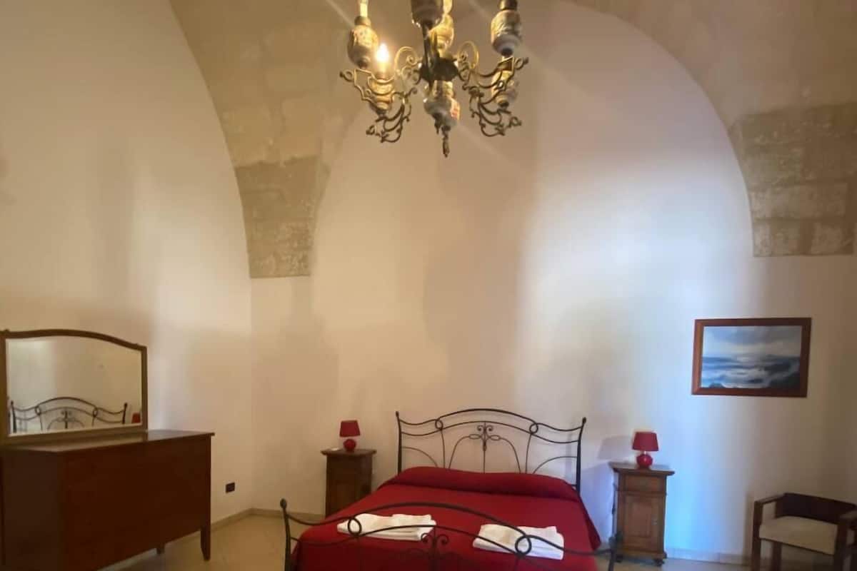 Un dormitorio con cama, cómoda, silla, mesita de noche, lámpara y un cuadro en la pared.