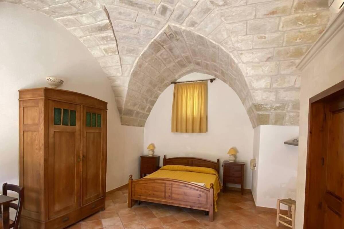 Un dormitorio con techo de piedra, un armario de madera, una cama con colcha amarilla y un taburete de madera.