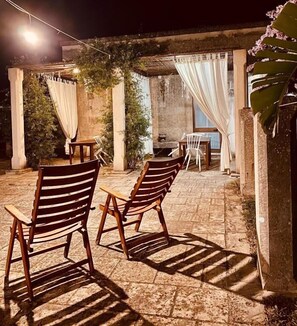 Un patio exterior con suelo de piedra, sillas de madera, una mesa y una lámpara colgante.