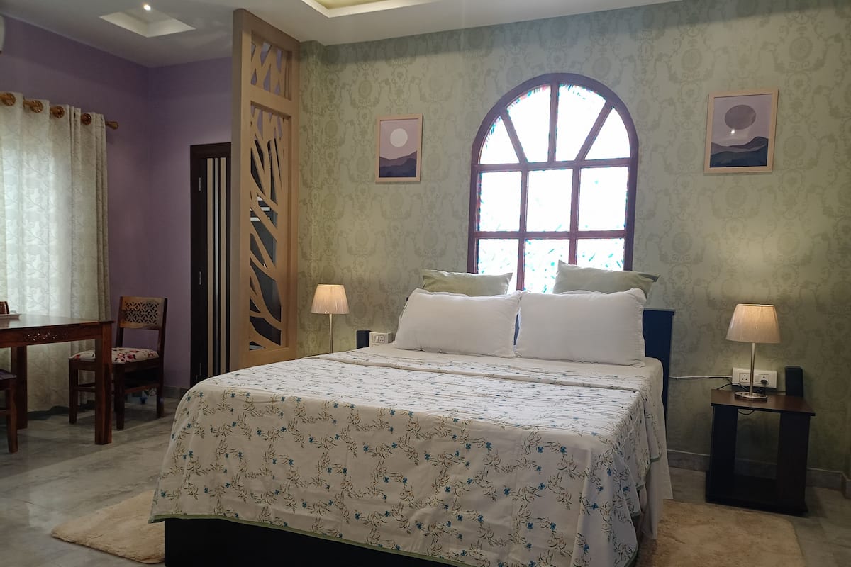 A bedroom with a large bed, two bedside tables with lamps, a chair, a desk, and a window with a view.