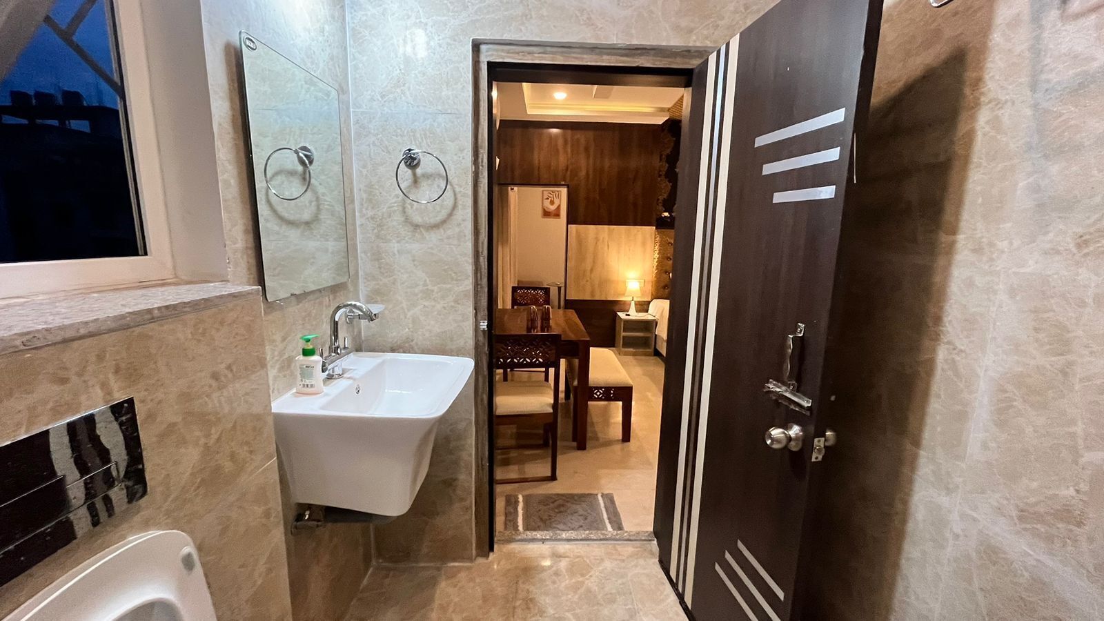 A bathroom with a sink, mirror, and towel rings. There is a door leading to a dining area with a table and chairs.