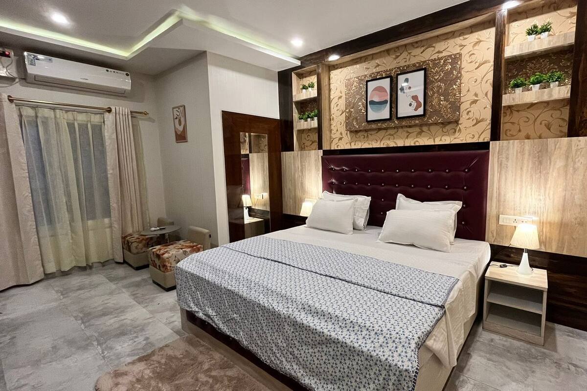 A modern hotel room with a large bed, a bedside table with a lamp, a decorative headboard, and a small seating area with a chair and a side table.
