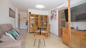 A living room with a sofa, a dining table with chairs, a wooden cabinet, and a television. - Oliva - Central apartment with air conditioning 150 meters from the beach in Mallorca (Alcúdia)