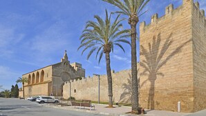 A historic building with arched windows and a large circular window, adjacent to a tall stone wall with battlements. - Oliva - Central apartment with air conditioning 150 meters from the beach in Mallorca (Alcúdia)