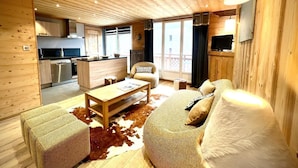 Interior acogedor de una cabaña con techo de madera, un comedor con mesa y sillas, un sofá con alfombra de cuero de vaca, y una cocina con electrodomésticos modernos.