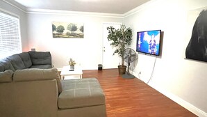 A living room with a sofa, a small table, a potted plant, a fan, and a TV mounted on the wall.