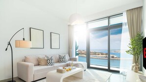 Una sala de estar moderna con sofá, mesa de centro, lámpara de pie y ventanas amplias que ofrecen vistas a un paisaje costero.