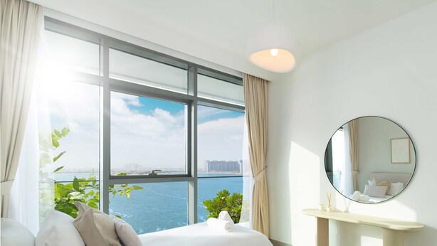 Habitación de hotel con un ventanal grande que ofrece vista a la ciudad, un espejo redondo en la pared y una mesita de noche con lámpara.