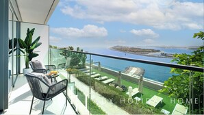 Un balcón con barandilla de vidrio que ofrece vistas a la costa, con una zona de estar que incluye una mesa y sillas, además de una planta en maceta.