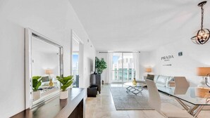 A modern living room with a glass table, a sofa, and a coffee table. There is a large mirror, a TV, and a plant. The room has a view of the city. - The Club 4320 (Miami)