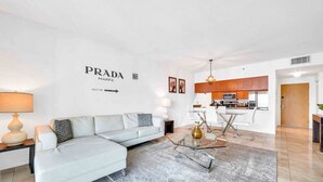 A modern living room with a white sofa, a glass coffee table, and a kitchen area in the background. - The Club 4320 (Miami)