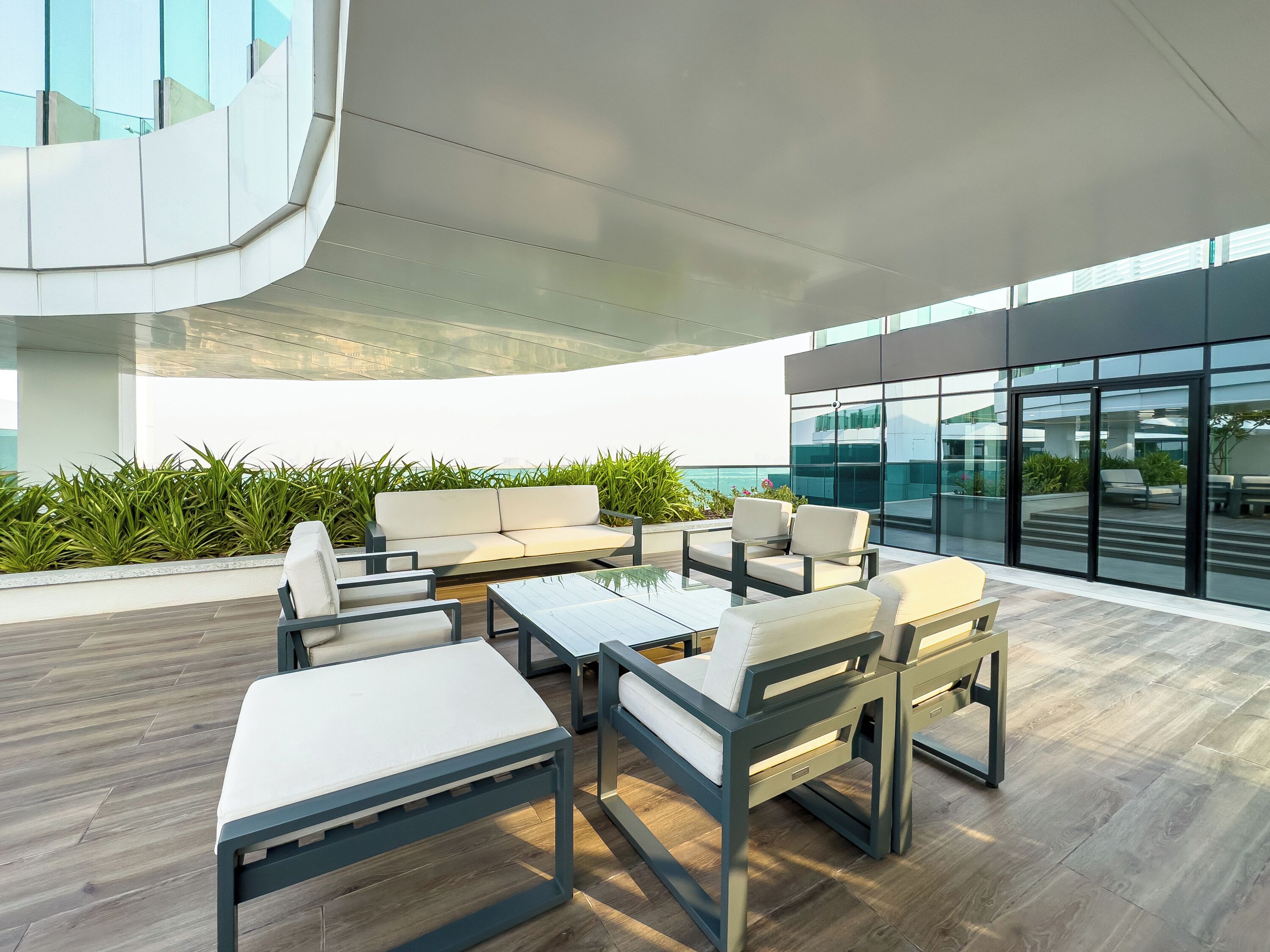 A modern outdoor seating area with a sofa and tables, surrounded by greenery and overlooking a cityscape.