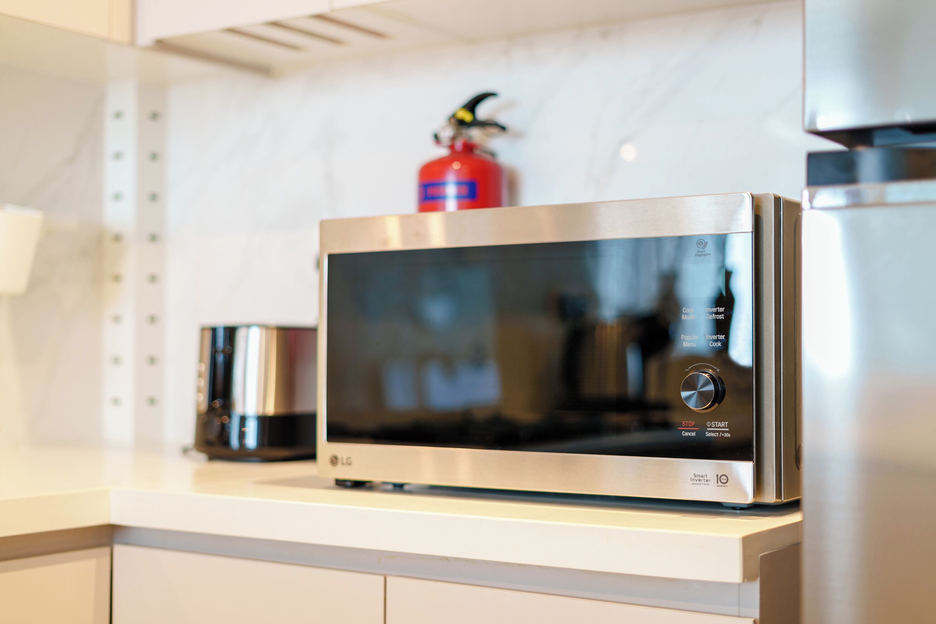 A modern kitchen with a stainless steel microwave on the counter, a red fire extinguisher mounted on the wall, and a stainless steel range hood above the counter.