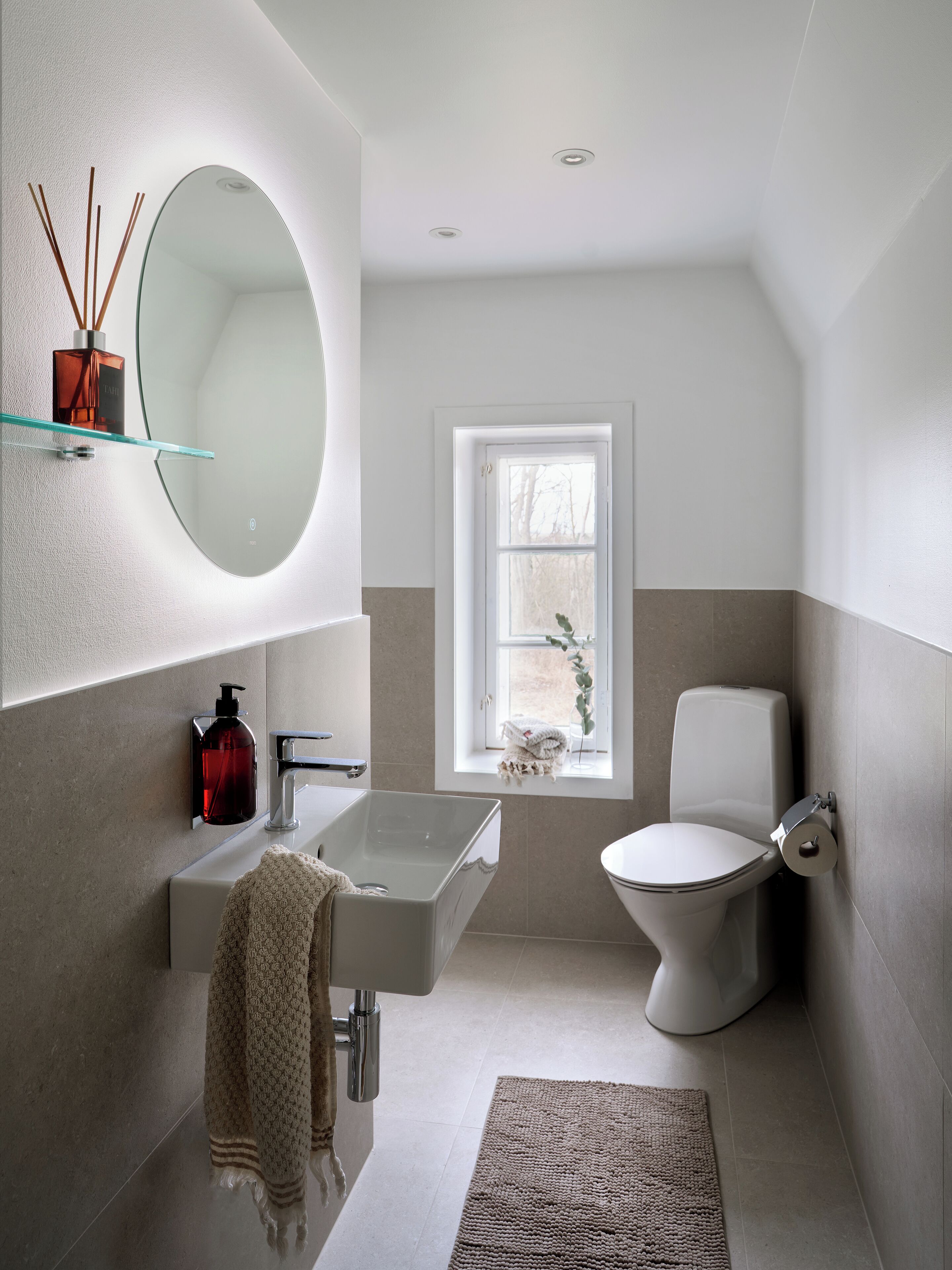 A modern bathroom with a glass sink, a round mirror, a toilet, and a window with a view of trees.