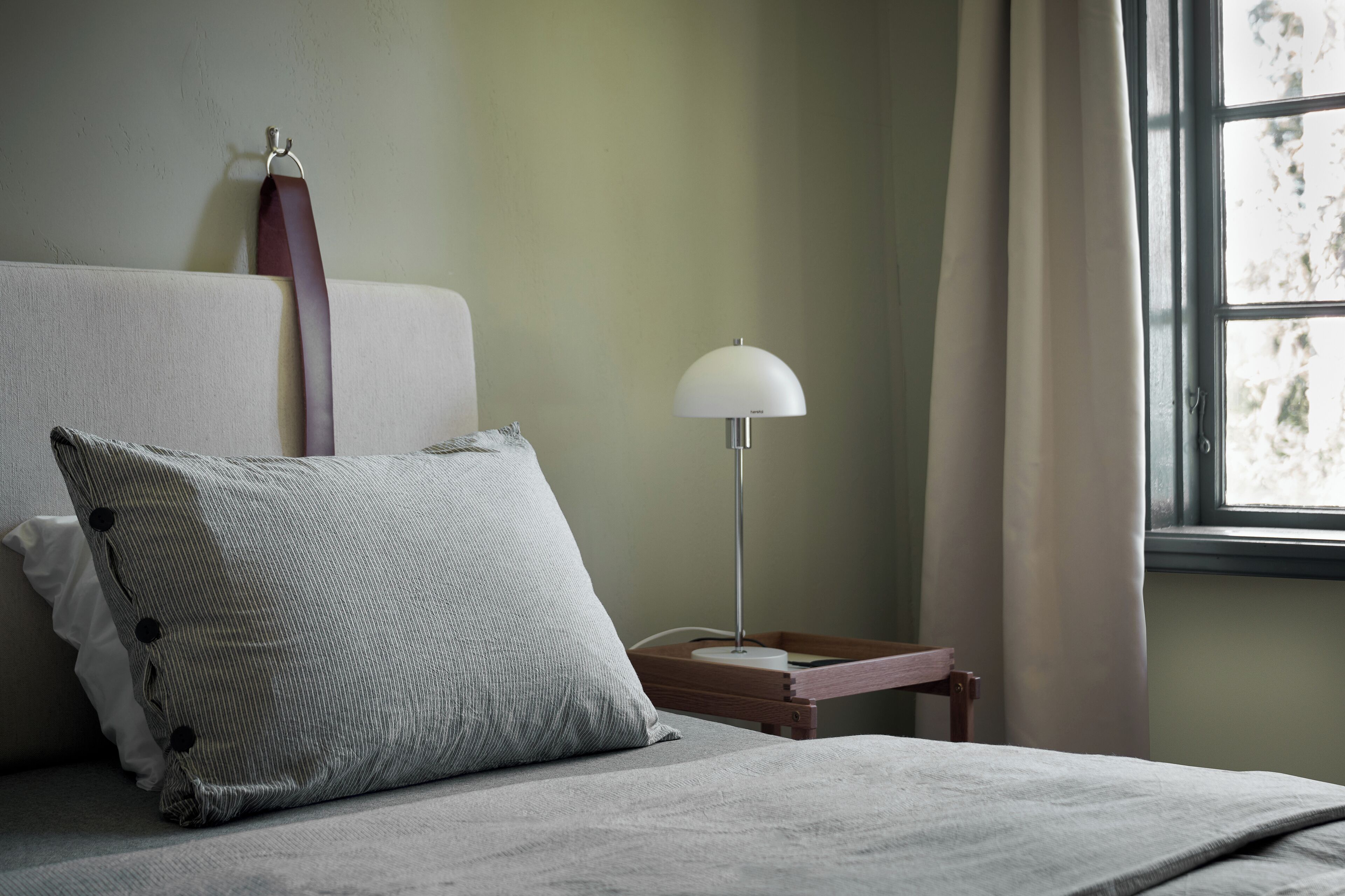 A neatly made bed with a grey pillow and a white pillow, a bedside table with a lamp, and a window with curtains.