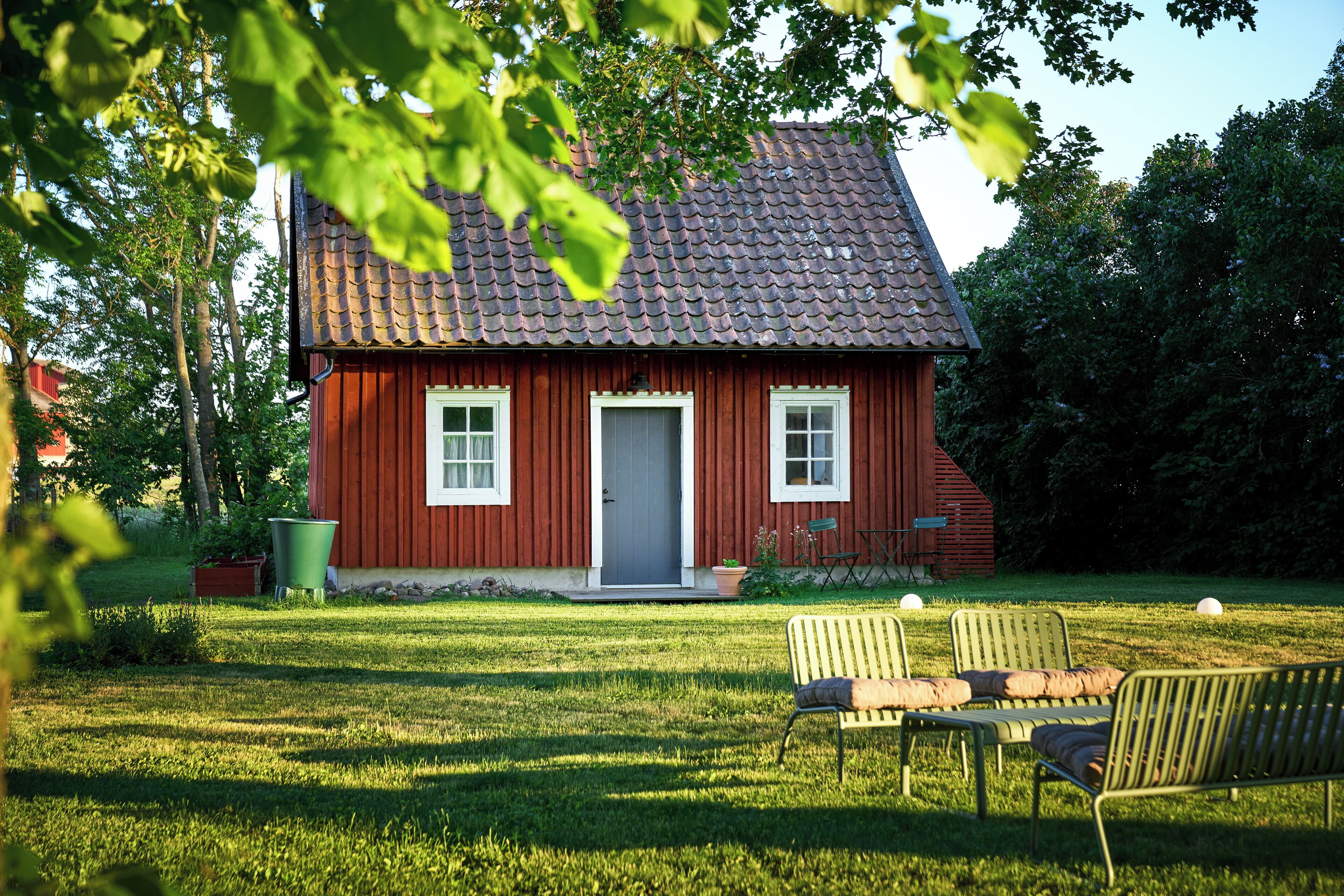 A red wooden house with a tiled roof, a front door, and two windows. There is a lawn with a bench and a trash can.