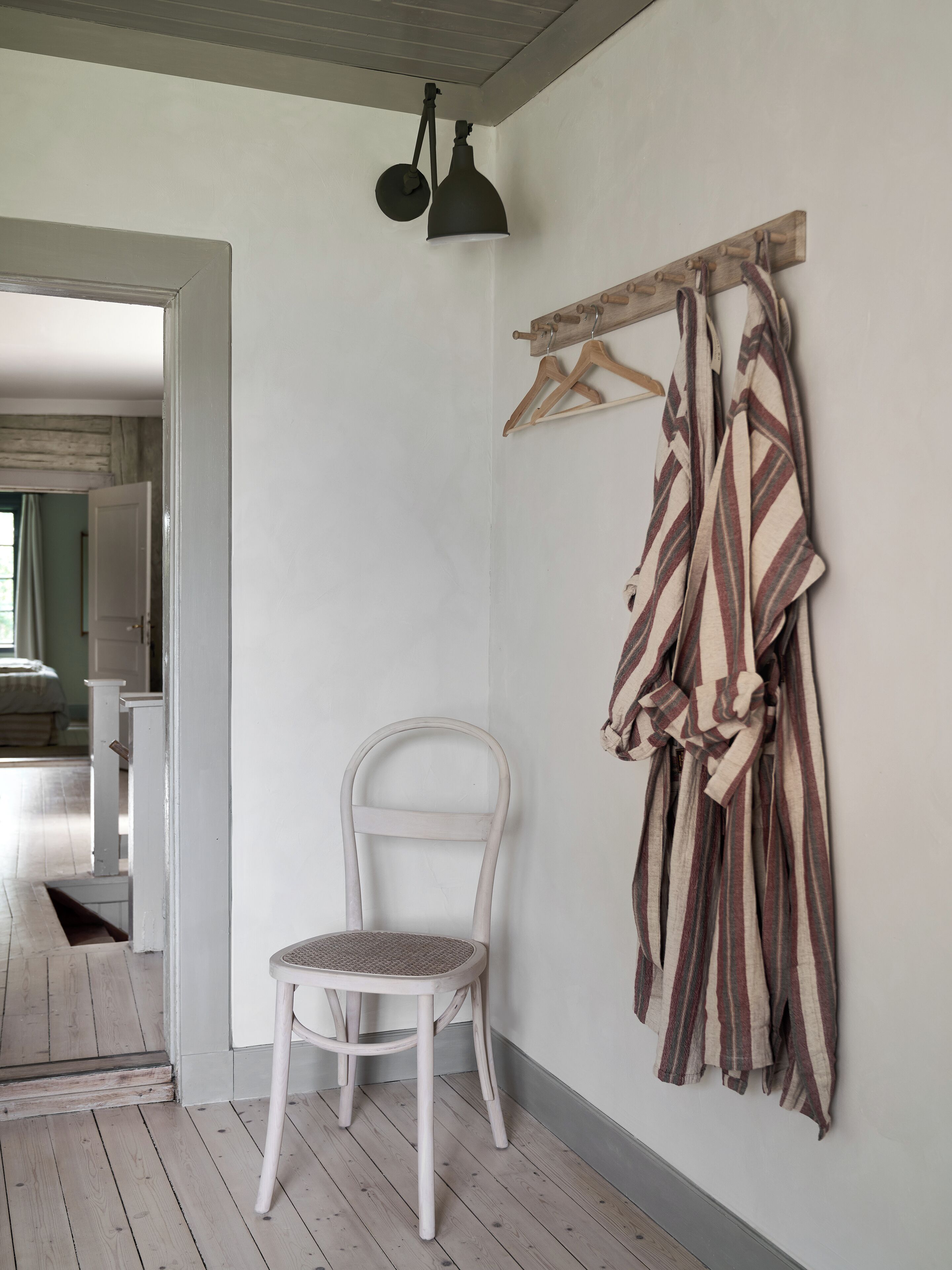A room with a white chair, a wooden coat rack with striped cloths, and a hanging light fixture.