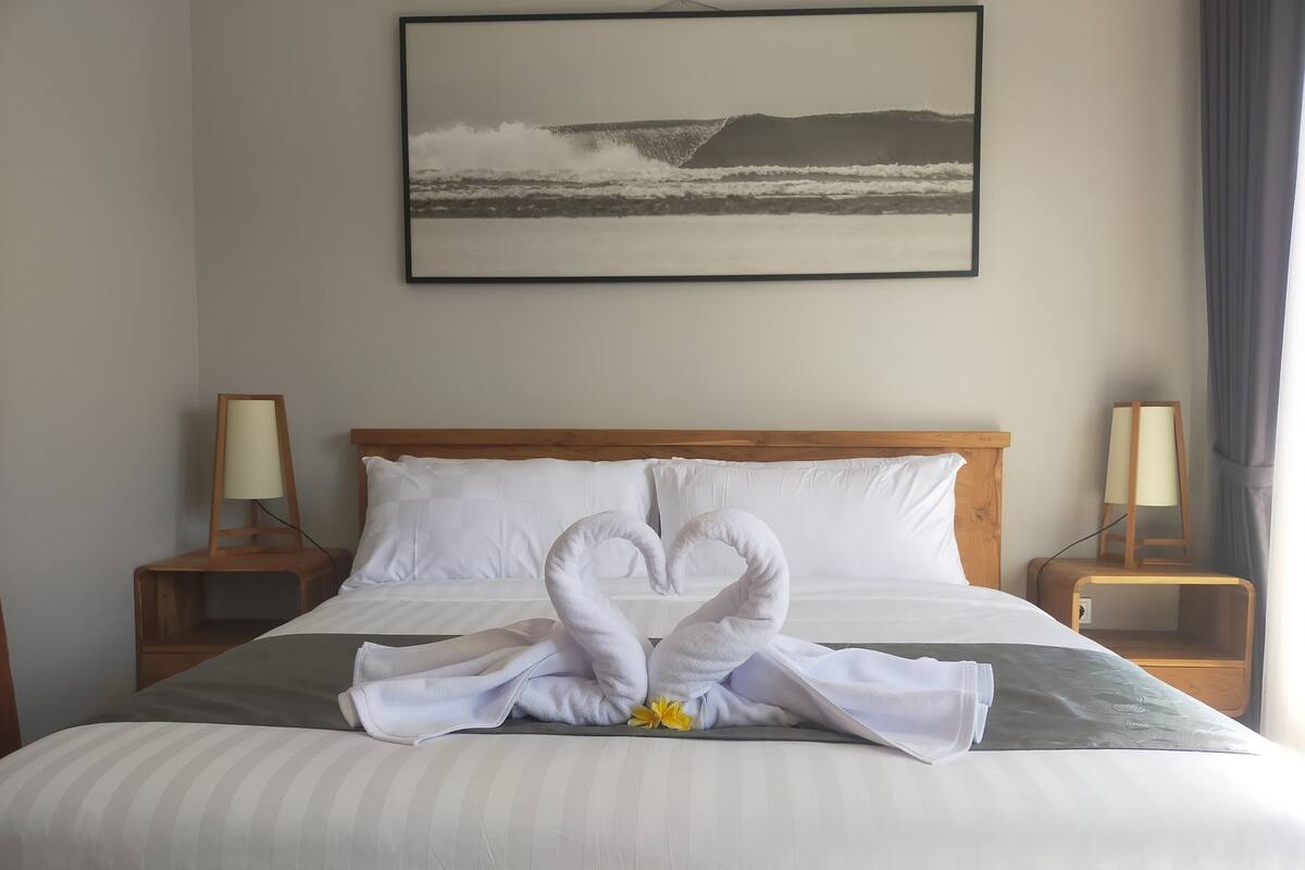 Een keurig opgemaakt bed met een handdoeksculptuur in het midden, een ingelijste foto aan de muur en nachtlampjes.