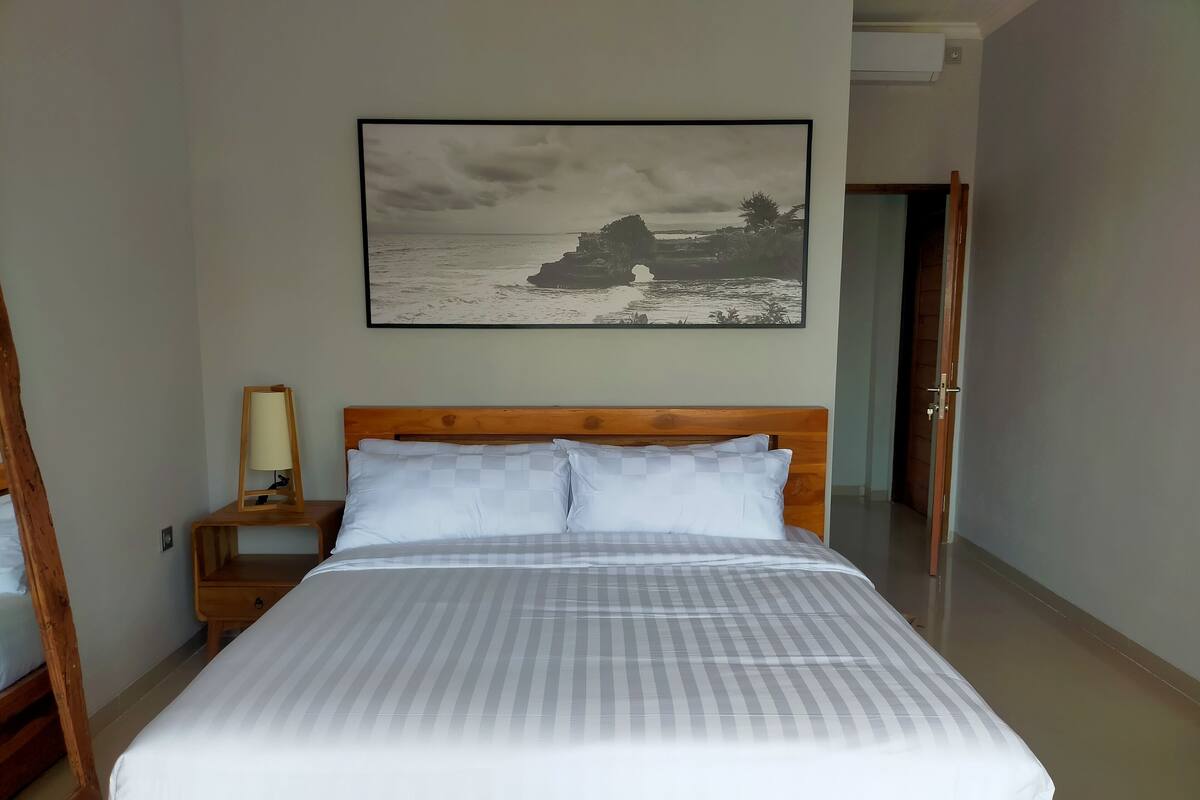 Una cama bien hecha con una colcha a rayas y dos almohadas, un cabecero de madera, una mesita de noche con una lámpara y un cuadro enmarcado de un paisaje costero en la pared.