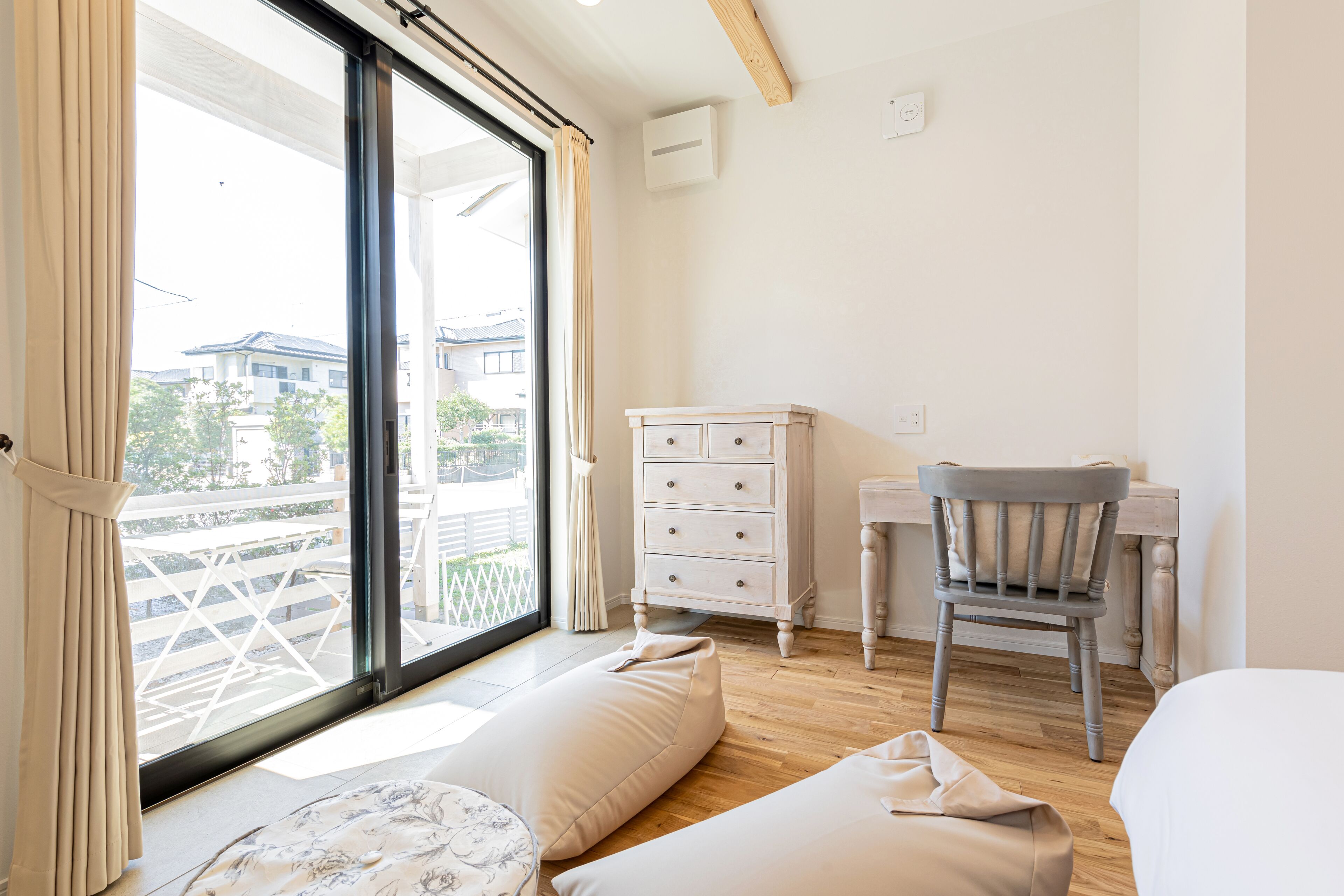 A room with a large window, a wooden desk, a chair, a bed, and a small table with a cushion.
