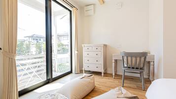 A room with a large window, a wooden desk, a chair, a bed, and a small table with a cushion.