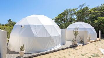 Dos tiendas blancas con forma de cúpula y una terraza de madera frente a ellas, rodeadas de plantas en macetas y árboles.