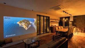 A modern living room with a large screen displaying a scenic island, wooden flooring, a sofa, a dining area with a table and chairs, and a kitchen with hanging lights. - Innosense Resort -In Chiba Resol- (Sanmu)