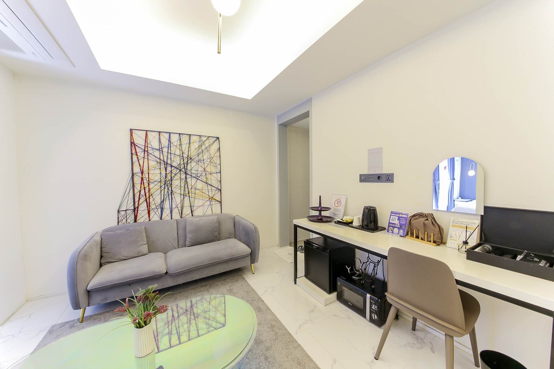 A modern hotel room with a grey sofa, a desk with a computer, a coffee maker, and a small table with a vase of flowers.