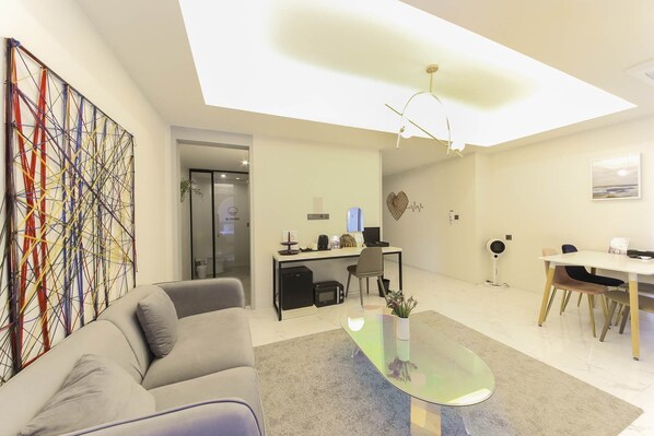 Una habitación de hotel moderna con una mesa de centro de vidrio, un sofá, un escritorio con una computadora y un cuadro abstracto grande en la pared.