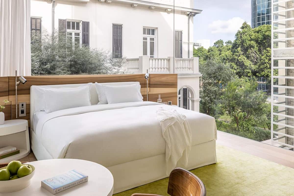 Habitación de hotel con una cama grande, una mesita de noche, una silla y vistas a zonas verdes y edificios.
